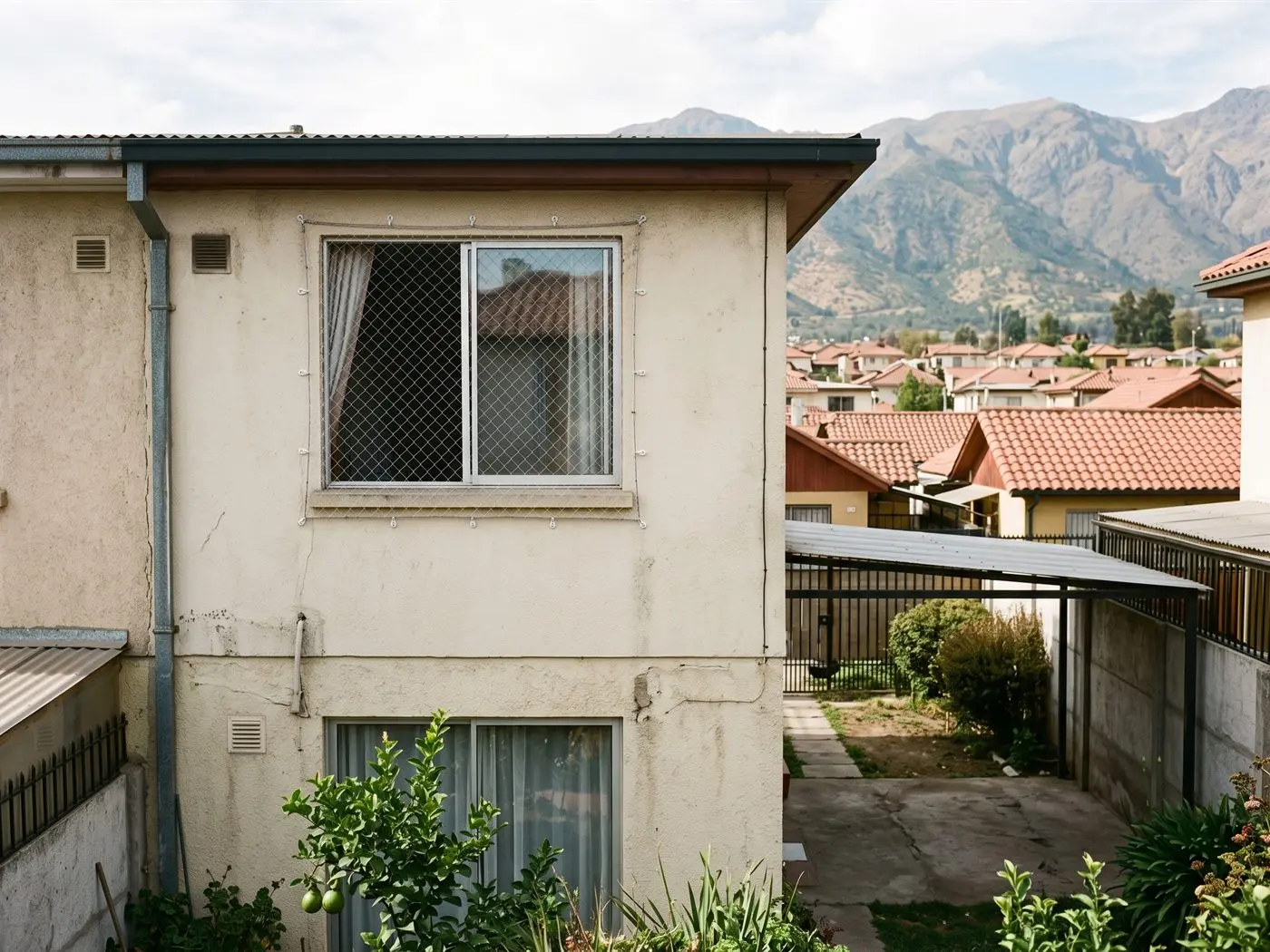 Ventana de segundo piso de una casa pareada de La Florida con malla de seguridad, con techos de tejas y la precordillera