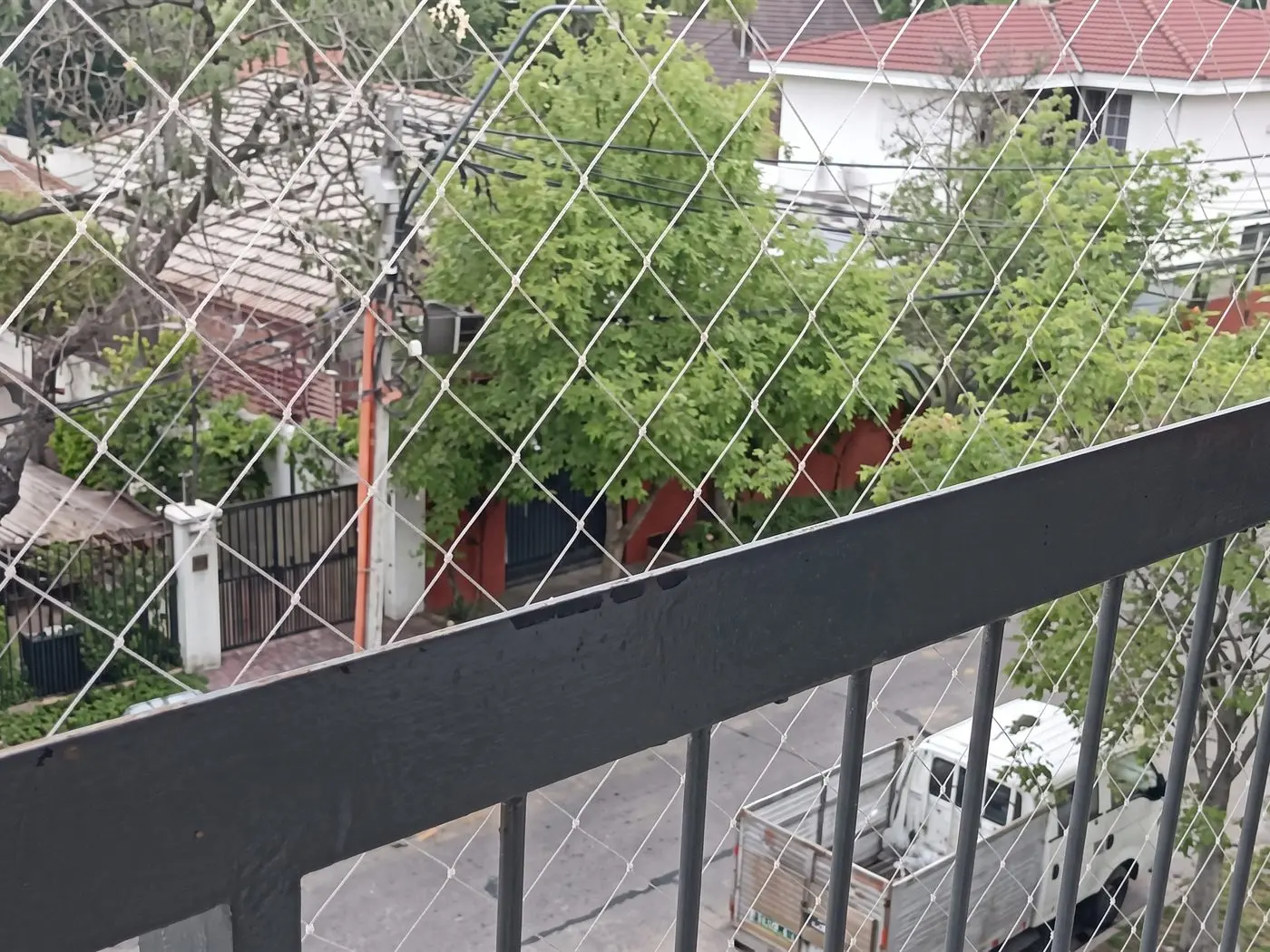 Malla de seguridad en el balcón de un edificio, con la baranda de fierro, una calle con árboles y una casa de techo rojo al fondo