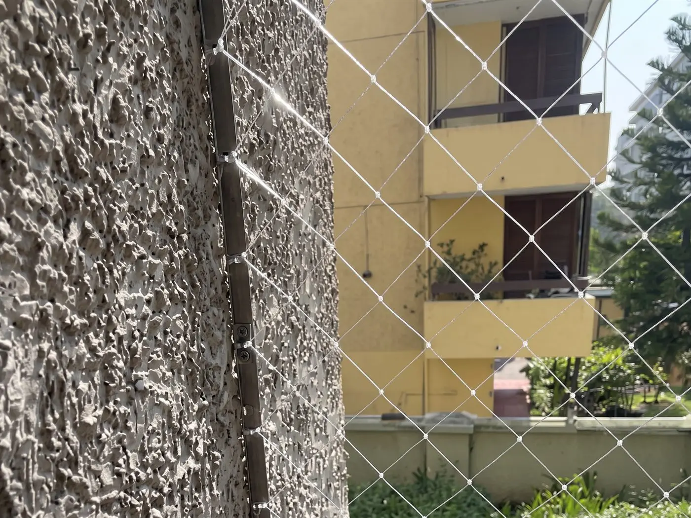 Malla de seguridad fijada junto a un pilar de hormigón, con un edificio amarillo detrás