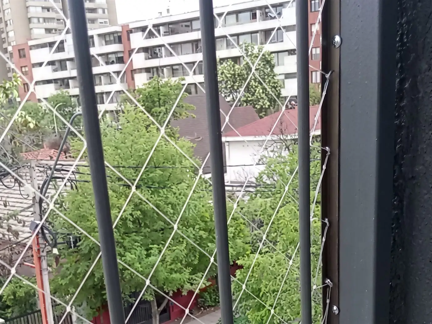 Malla de seguridad fijada con ganchos al borde de un pilar de fierro, junto a los barrotes de la baranda, con un edificio de departamentos y árboles detrás