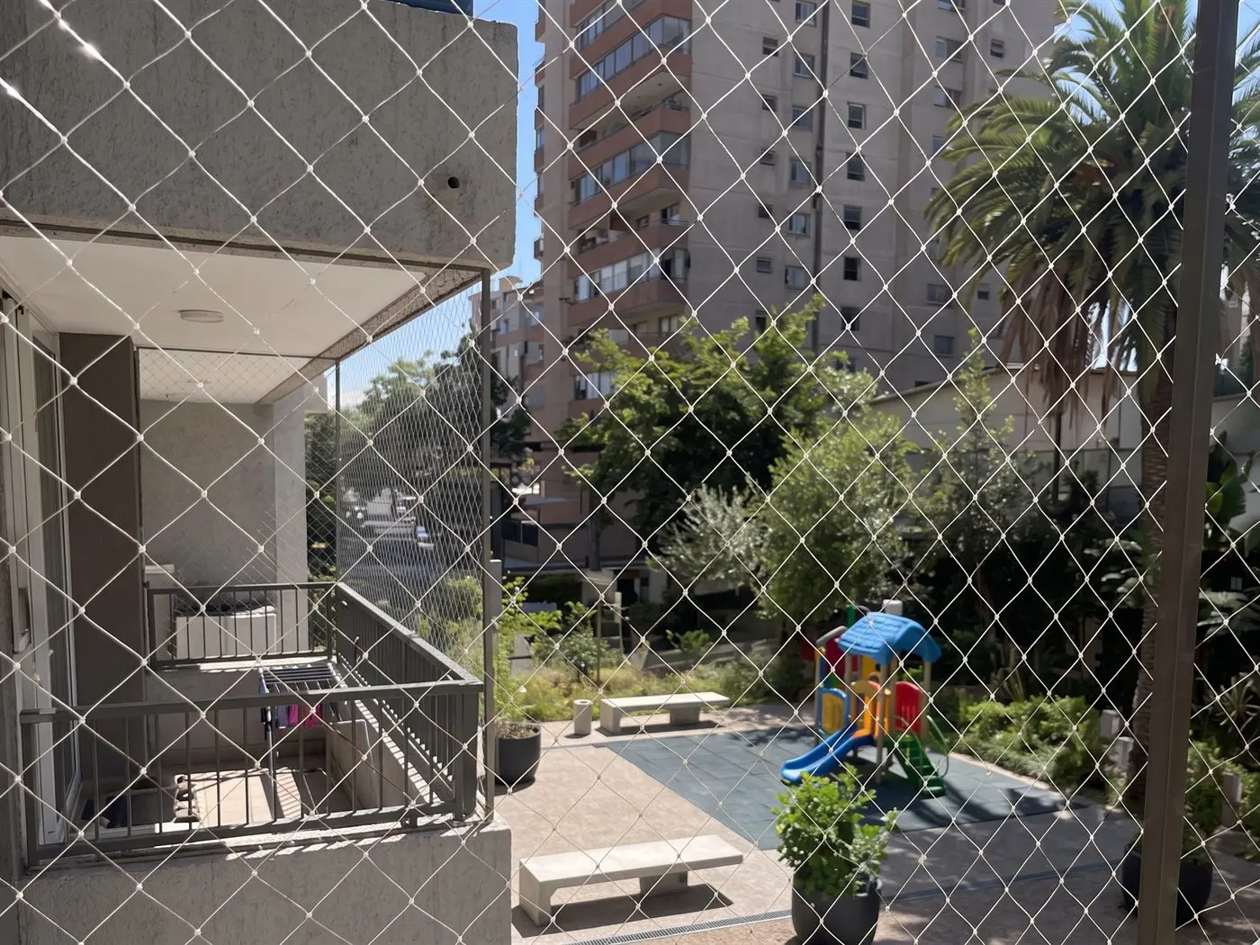 Malla de seguridad en un balcón de condominio, con el patio común, los juegos y la torre vecina detrás